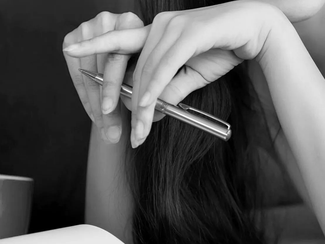 Verschränkte Hände mit silbernem Kugelschreiber vor langem dunklem Haar, darunter ein aufgeschlagenes Heft